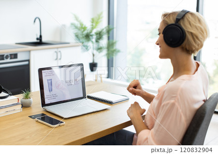 Happy Caucasian woman orders cosmetics online using laptop while listening to music in headphones. She sits in modern home office setting with smartphone and notebook on desk. 120832904