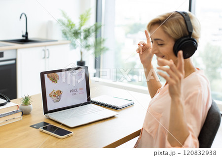 Happy Caucasian woman ordering food online through delivery website using laptop while listening to music in headphones. She sits at home workspace, excited and relaxed with headphones on. 120832938