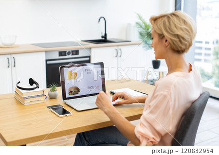 Happy Caucasian woman ordering food online using laptop in bright modern kitchen. She sits at wooden table with smartphone and notebook. Digital menu on screen displays food choices. 120832954