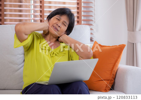 Asian senior woman sitting on sofa with headache while using laptop computer in living room. Asian senior woman sitting on sofa with headache while using laptop computer in living room. 120833113
