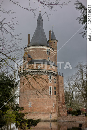 A historic brick castle with tall spire-like towers reflected in a calm moat, surrounded by leafless trees on a cloudy day, creating a serene and mysterious atmosphere. Castle Duurstede. 120836033