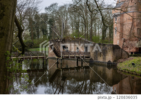 A serene medieval scene of a ruined brick wall, wooden drawbridge, and calm moat, surrounded by bare trees and reflected in the water under a cloudy sky. Castle Duurstede. A serene medieval scene of a ruined brick wall, wooden drawbridge, and calm moat, surrounded by bare trees and reflected in the water under a cloudy sky. Castle Duurstede. 120836035
