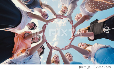 Young women graduates celebrate success outdoors on a sunny day, forming a circle with hands raised in unity, smiling and looking up at the blue sky 120839689