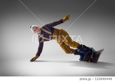 Snowboarder wearing mustard pants and knit hat leans gracefully mid-motion on his snowboard against gray studio backdrop. 120840446