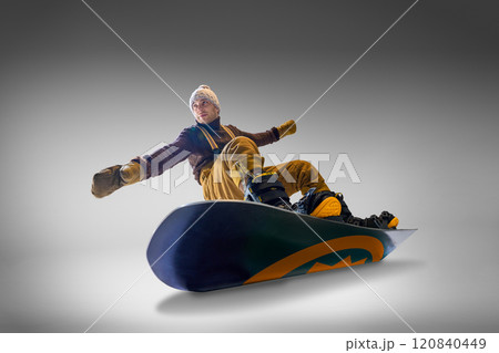 Snowboarder dressed in brown sweater and mustard pants leans back on his snowboard against gray background, showcasing balance and skill. 120840449