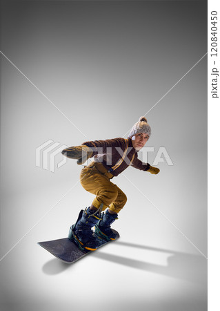 Snowboarder wearing cozy knitted sweater and gloves, mustard pants demonstrating stylish balance against grey studio background. 120840450