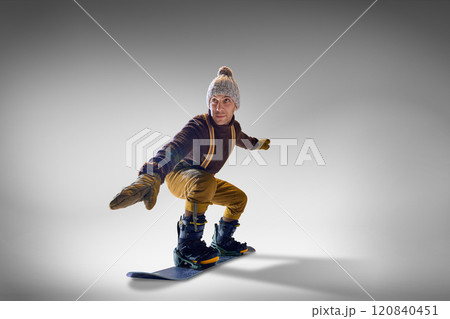 Snowboarder wearing cozy knitted sweater and gloves, mustard pants demonstrating stylish balance against grey studio background. 120840451