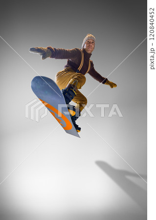 Snowboarder in brown sweater and mustard pants showcasing balance on his snowboard against grey studio background. 120840452