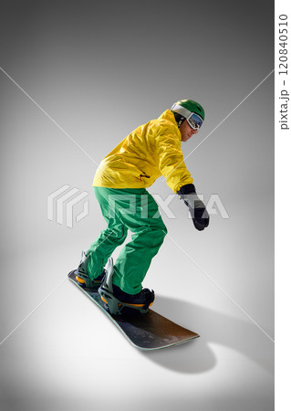 Side view, full length photo of athletic young man elegantly slopes on snowboard in motion, dressed vibrant clothes against grey studio background. 120840510