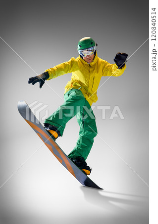 Young athletic man dressed modern, bright yellow and green snowboarding clothes performing tricks in motion against grey studio background. Young athletic man dressed modern, bright yellow and green snowboarding clothes performing tricks in motion against grey studio background. 120840514