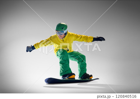 Athletic snowboarder dressed modern winter outfit snowboarding, performing tricks on snowboard against grey studio background. 120840516