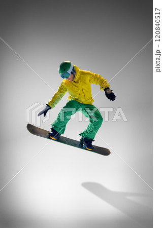 Young athletic man dressed vibrant yellow and green winter gear, performing tricks on snowboard against grey studio background. 120840517