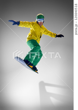 Young athletic man dressed vibrant yellow and green winter gear, performing tricks on snowboard against grey studio background. 120840518