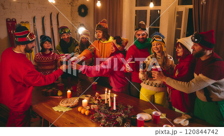 Friends in colorful winter attire stand around rustic wooden table, toasting with red cups while candles, oranges, and pastries create festive atmosphere. Friends in colorful winter attire stand around rustic wooden table, toasting with red cups while candles, oranges, and pastries create festive atmosphere. 120844742