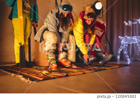 Two friends in colorful ski gear sit on bench indoors, fastening their ski boots, surrounded by cozy festive atmosphere with winter decorations. 120845881