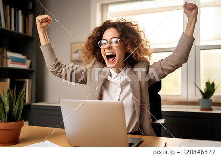 Excited and joyful young businesswoman at her desk with a laptop, celebrating a successful 120846327