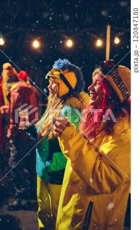 Friends in bright winter outfits celebrate outdoors with drinks, smiling and laughing as snow falls, creating festive holiday atmosphere. Friends in bright winter outfits celebrate outdoors with drinks, smiling and laughing as snow falls, creating festive holiday atmosphere. 120847180