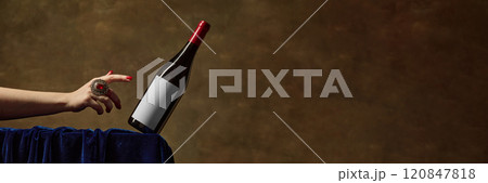 Mockup showing red wine bottle with blank label, being gracefully balanced by hand adorned with decorative red-stoned ring, against rich brown textured background. 120847818