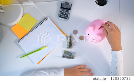 High angle view of young girl placing coins and banknotes into a piggy bank while seated at a bright organized table filled with stationery and a calculator managing personal finances High angle view of young girl placing coins and banknotes into a piggy bank while seated at a bright organized table filled with stationery and a calculator managing personal finances 120850753