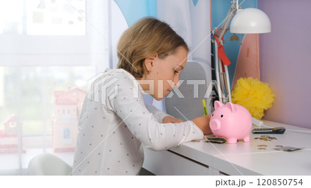 Young girl diligently places coins into a pink piggy bank while taking notes about her savings. Personal finance and money management Young girl diligently places coins into a pink piggy bank while taking notes about her savings. Personal finance and money management 120850754