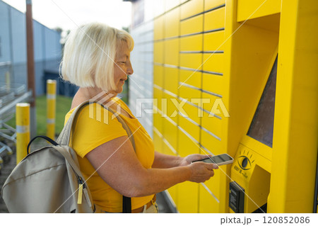Happy smiling 50s senior woman client with smart phone at outdoor automated parcel machine hoosing operation he touch screened on smart background Happy smiling 50s senior woman client with smart phone at outdoor automated parcel machine hoosing operation he touch screened on smart background 120852086