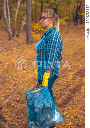 A tired female volunteer after cleaning up garbage in the forest stands with a garbage bag in her hands and looks sadly to the side. A woman worries about food and its pollution by garbage waste. 120852113