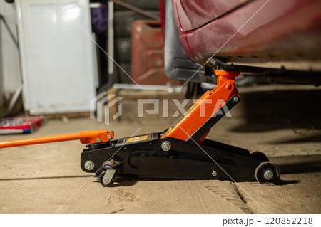 The car is standing in the garage, raised with a jack, while a man is repairing it Close-up of a jack under the bottom of a car in a garage. The jack holds the car while the person changes the wheel. 120852218