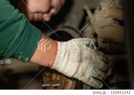 A man in work gloves repairs a wheel on a car in a garage. A man replaces a broken part on his car in the garage in the evening. Self-repair machine concept. A man in work gloves repairs a wheel on a car in a garage. A man replaces a broken part on his car in the garage in the evening. Self-repair machine concept. 120852242