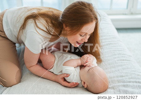 Happy family at home. Mother playing with little toddler child on bed. Mom baby relax playing having fun together. Mother looks at baby with love care. Mom of breast feeding baby rest in living room 120852927