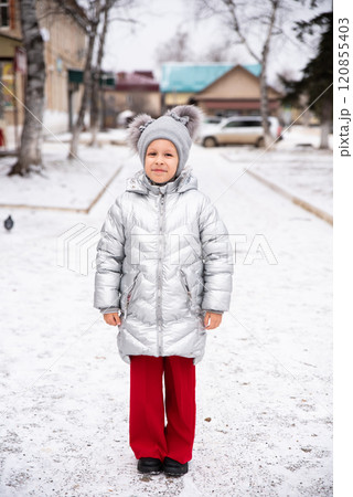 8-year-old Russian girl smiles in the snow, wearing a warm winter jacket and hat. She stands outdoors in a snowy landscape, looking at the camera with a joyful, playful expression 120855403