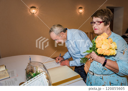 An elderly couple signs documents at a registry office in Russia, celebrating their 50th wedding anniversary. The woman holds a bouquet of yellow roses, symbolizing love and commitment 120855605