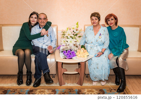76-year-old man and 71-year-old woman celebrate their golden wedding anniversary, sitting on a sofa in the registry office, embraced by their daughter and granddaughter, holding a bouquet and medal 76-year-old man and 71-year-old woman celebrate their golden wedding anniversary, sitting on a sofa in the registry office, embraced by their daughter and granddaughter, holding a bouquet and medal 120855616