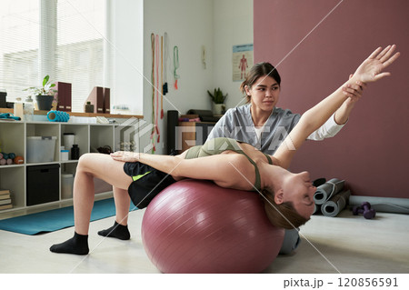 Assisting Woman with Stretching Over Exercise Ball 120856591