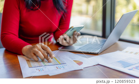Woman wear a red shirt holding a pen and smartphone while working at office, Close up shot hands of woman use pen to analyze graphs and business data. Woman wear a red shirt holding a pen and smartphone while working at office, Close up shot hands of woman use pen to analyze graphs and business data. 120859323