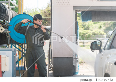 高圧洗浄機の洗車ガンを使い洗車するつなぎを着た整備士の男性 120859718