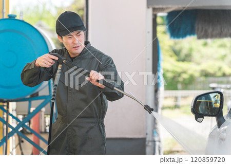 高圧洗浄機の洗車ガンを使い洗車するつなぎを着た整備士の男性 120859720
