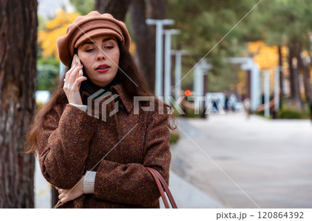 Mid shot of a young Caucasian woman in a hat and coat talking on phone. Modern communication and business. Copy space Mid shot of a young Caucasian woman in a hat and coat talking on phone. Modern communication and business. Copy space 120864392