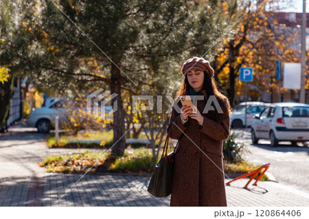 Wide shot of a young Caucasian woman in a autumn coat and beret using smartphone and walking on autumn street. Modern communication and business 120864406