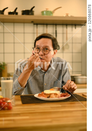 Happy middle aged man enjoys a homemade breakfast at wooden kitchen counter Happy middle aged man enjoys a homemade breakfast at wooden kitchen counter 120868078