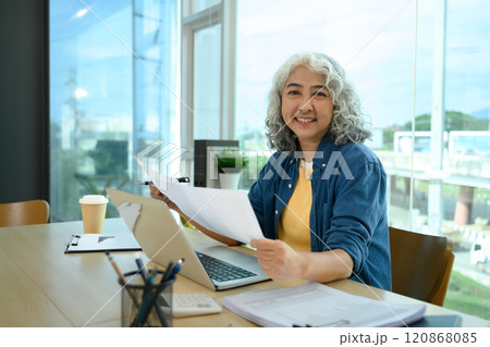 Confident senior businesswoman reviewing paperwork at her desk in modern office 120868085