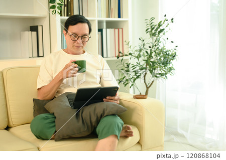 Happy middle aged man sipping coffee from a green mug and browsing on digital tablet Happy middle aged man sipping coffee from a green mug and browsing on digital tablet 120868104