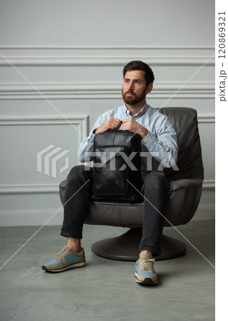A man is comfortably sitting in a chair, resting with a leather backpack near him 120869321