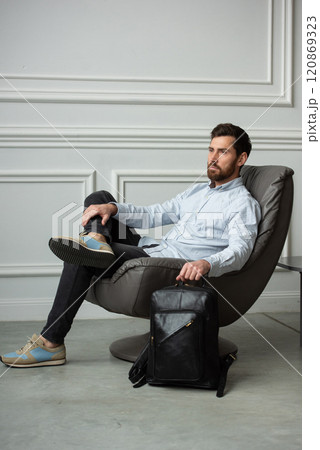 A man is comfortably sitting in a chair, resting with a leather backpack near him 120869323