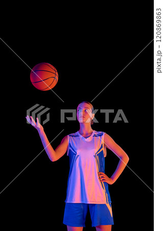 Portrait of young girl in uniform, basketball player bouncing ball on hand against black studio background in neon light 120869863