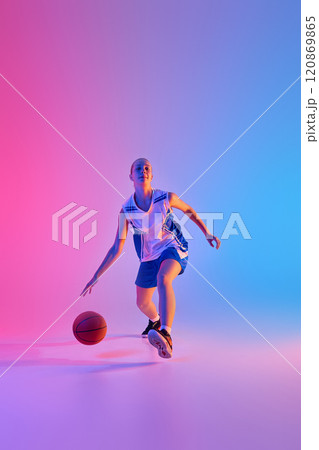 Full-length dynamic image of teen girl in uniform, basketball player in motion with ball during training session against gradient pink blue background in neon light 120869865