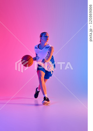 Full-length dynamic image of teen girl in uniform, basketball player in motion with ball during training session against gradient pink blue background in neon light 120869866