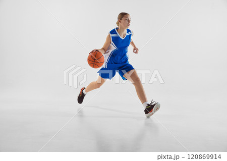 Young teen girl in blue uniform, in motion with ball, playing basketball, training against white studio background 120869914