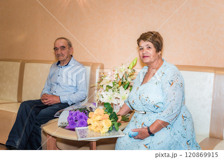 A senior Russian couple, 71 and 75 years old, just married at the registry office. They sit beside floral bouquets, both in blue attire, symbolizing family values and love A senior Russian couple, 71 and 75 years old, just married at the registry office. They sit beside floral bouquets, both in blue attire, symbolizing family values and love 120869915
