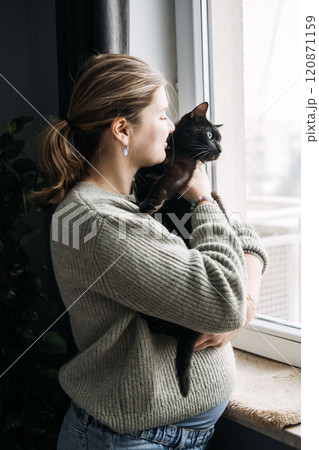 Pregnant woman holding black cat by the window, questioning the safety of keeping pets during pregnancy. Cozy home with coffee and candle. 120871159