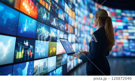 壁のモニター画面をチェックしながらノートパソコンで働くスーツ姿の女性 120872040